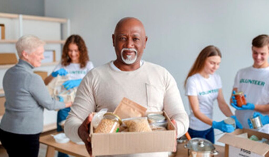 A black man with silver goatee holds a box of food at a food drive